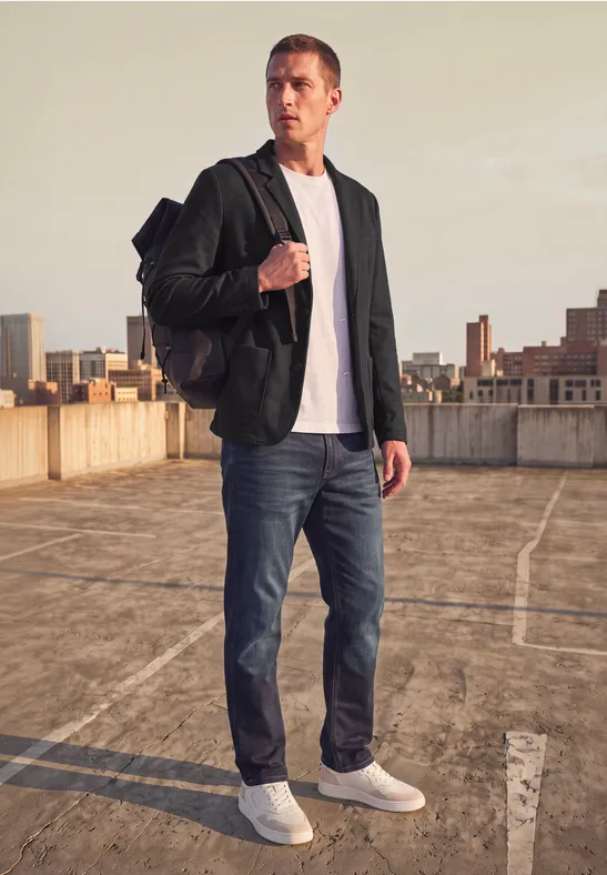 Ein junger Mann mit Rucksack, Blazer und Jeans steht auf einem Dachparkplatz, Blick auf die Stadt im Hintergrund.