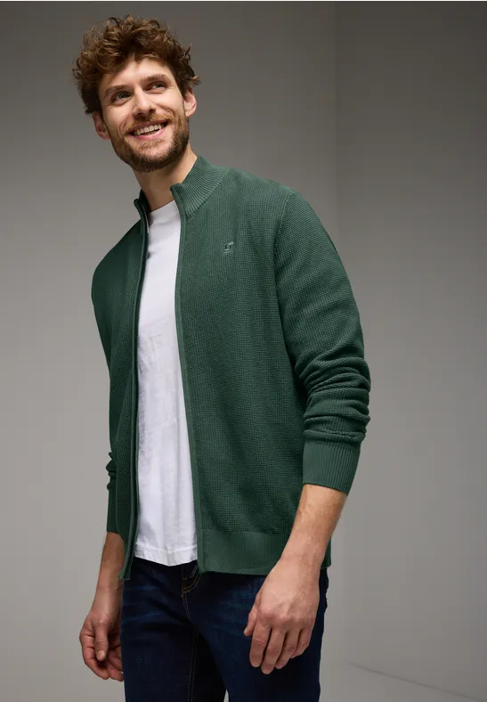 Homme souriant portant un cardigan vert et un t-shirt blanc.