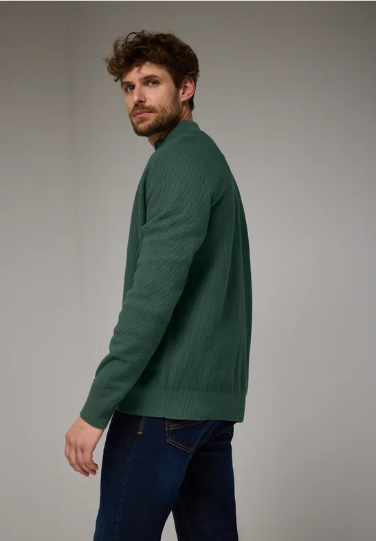 Homme en pull vert et jean regardant sur le côté.