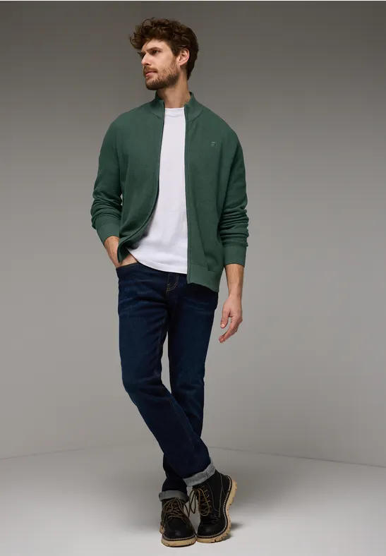 Homme debout, veste verte, t-shirt blanc et jean bleu.