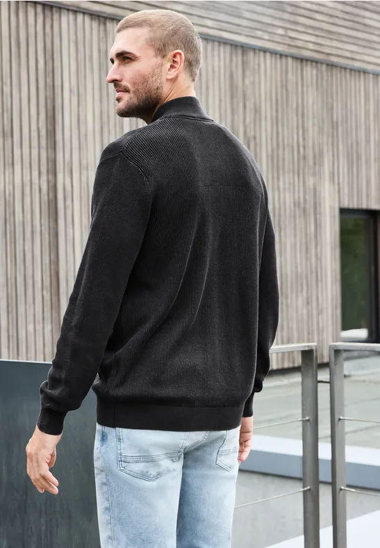 Mann mit schwarzem Pullover und hellen Jeans steht seitlich auf einer Terrasse vor modernem Gebäude.