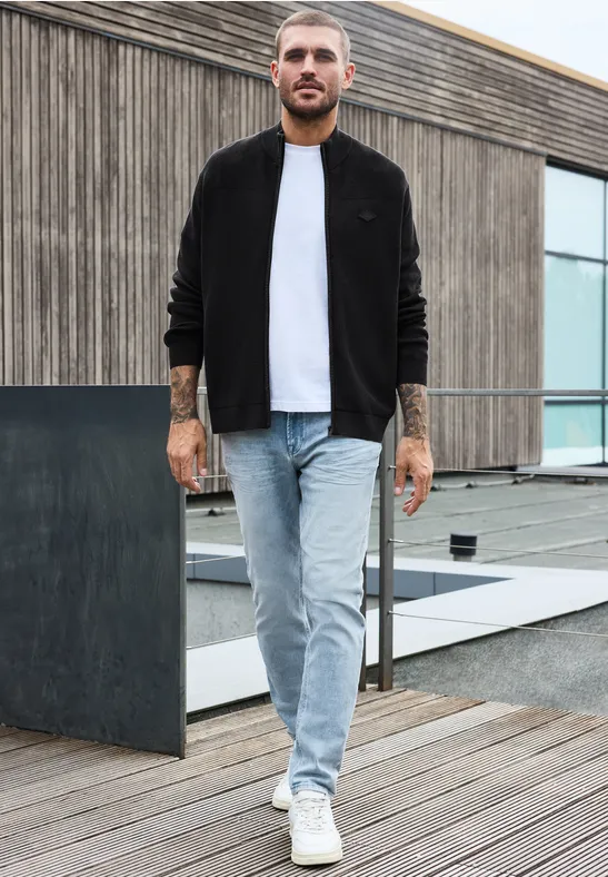 Ein bärtiger Mann in schwarzer Jacke, weißem T-Shirt und Jeans steht auf einer Holzterrasse vor modernem Gebäude.