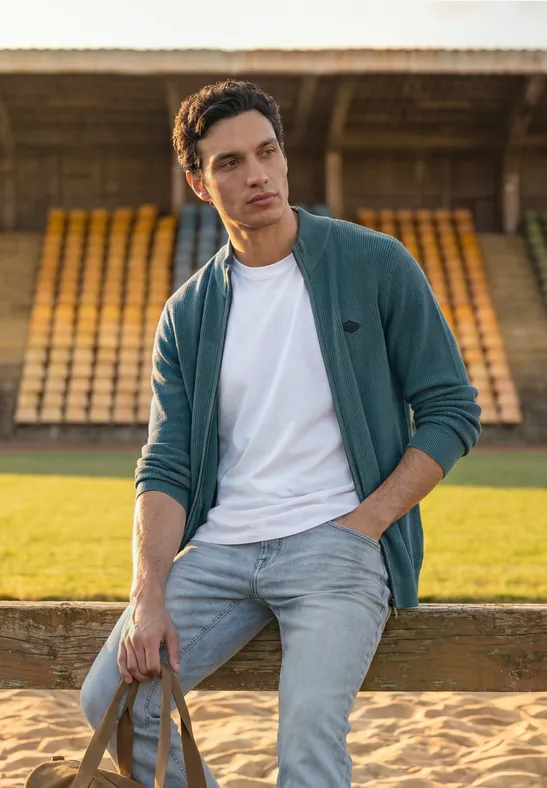 Ein junger Mann in Jeans und Jacke lehnt mit Tasche vor leeren Tribünen auf einem Sportplatz.