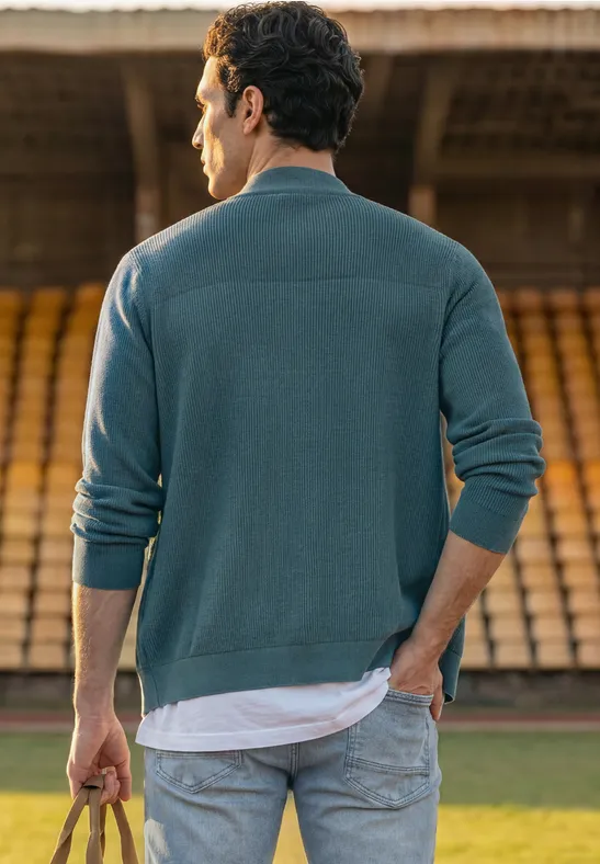 Ein Mann steht mit dem Rücken zur Kamera in einem Stadion, trägt Jeans, Pullover und hält einen Fußball.