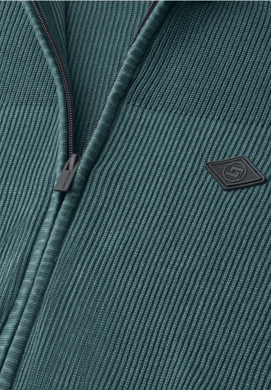 Nahaufnahme einer dunkelgrünen, gerippten Strickjacke mit Reißverschluss und kleinem Logopatch auf der Brust.