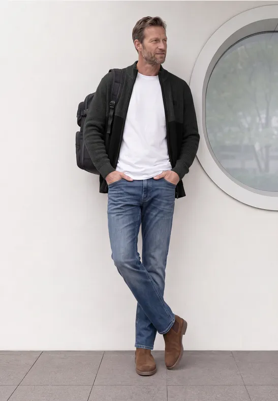 Een nonchalant geklede man leunt tegen een muur bij een rond raam, draagt jeans, wit shirt, jas en rugzak.