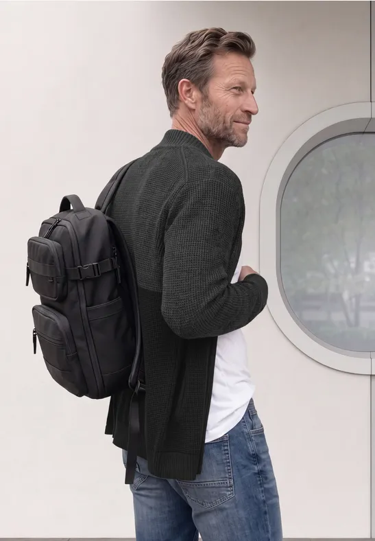 Een man in jeans en colbert draagt een zwarte rugzak en kijkt opzij voor een witte muur met een rond raam.