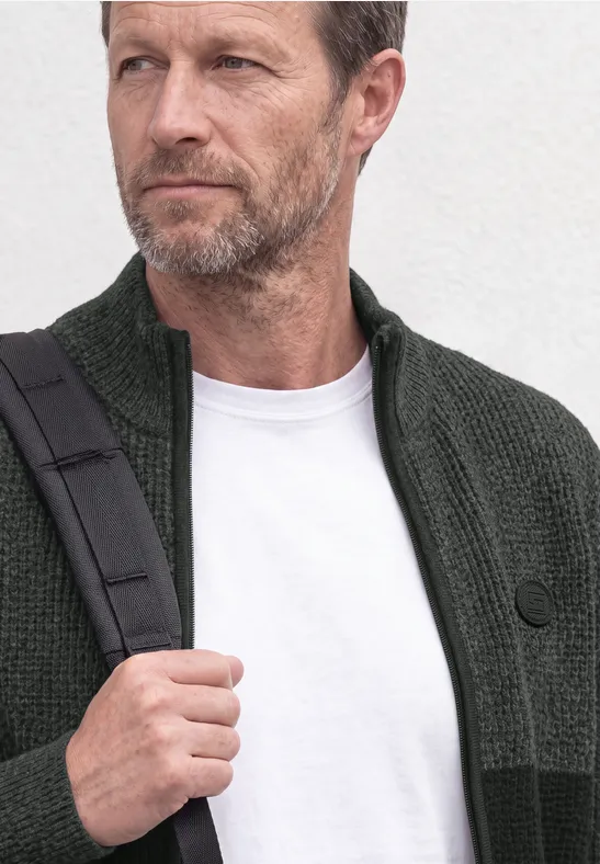 Een man van middelbare leeftijd met grijze baard draagt een groen vest, wit T-shirt en schoudertas.