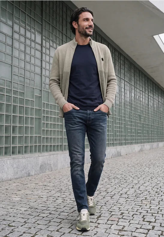 Een man met baard in een beige jas, donkerblauw T-shirt en spijkerbroek loopt over een geplaveid trottoir.