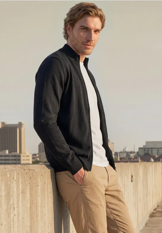 Een man met zwarte jas, wit T-shirt en beige broek leunt ontspannen tegen een dakrand met stadszicht.