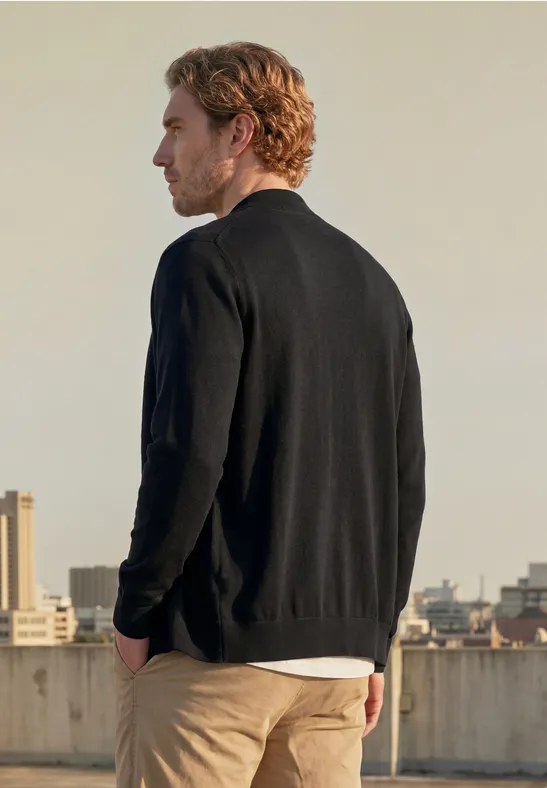 Een man in een zwarte trui en beige broek staat op een dak, met stadsgebouwen op de achtergrond.