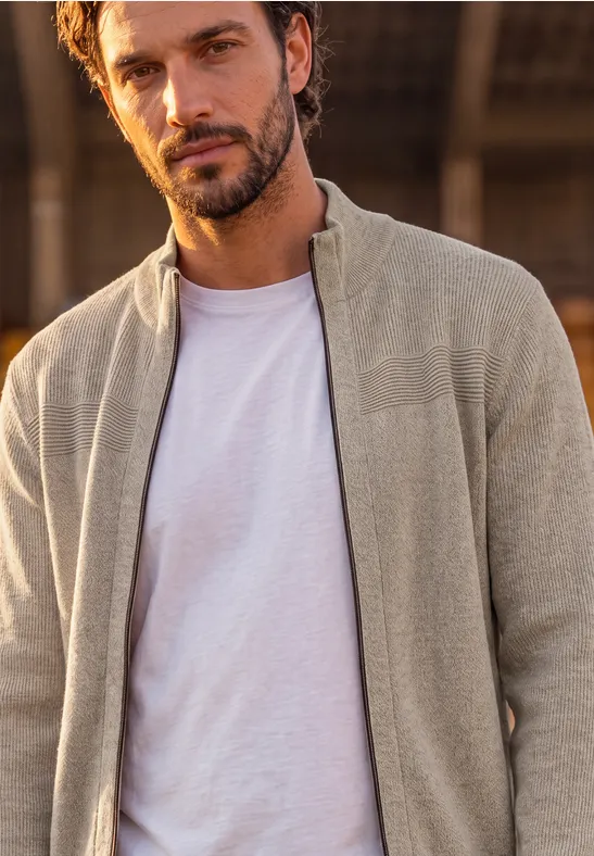 Ein gutaussehender Mann mit Bart trägt ein weißes T-Shirt und eine beige Strickjacke vor unscharfem Hintergrund.