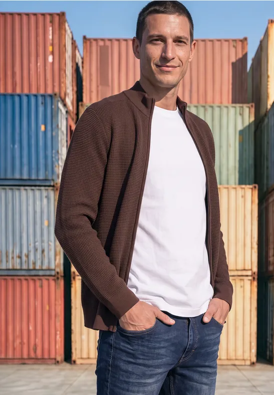 Ein Mann in Jeans, weißem T-Shirt und brauner Jacke steht lächelnd vor bunten Containern.