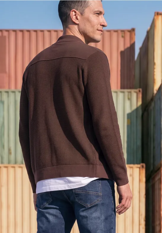 Ein Mann in brauner Jacke und Jeans steht vor bunten Frachtcontainern und blickt weg.