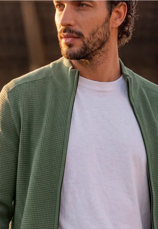Ein Mann trägt eine grüne, strukturierte Jacke mit Reißverschluss über einem weißen T-Shirt.
