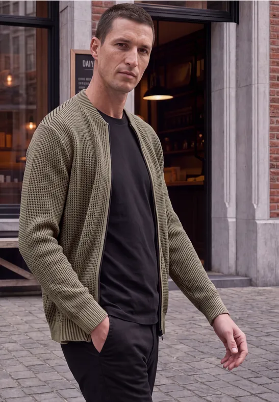 Ein Mann in grüner Strickjacke und schwarzem T-Shirt läuft vor einem Café in der Stadt.