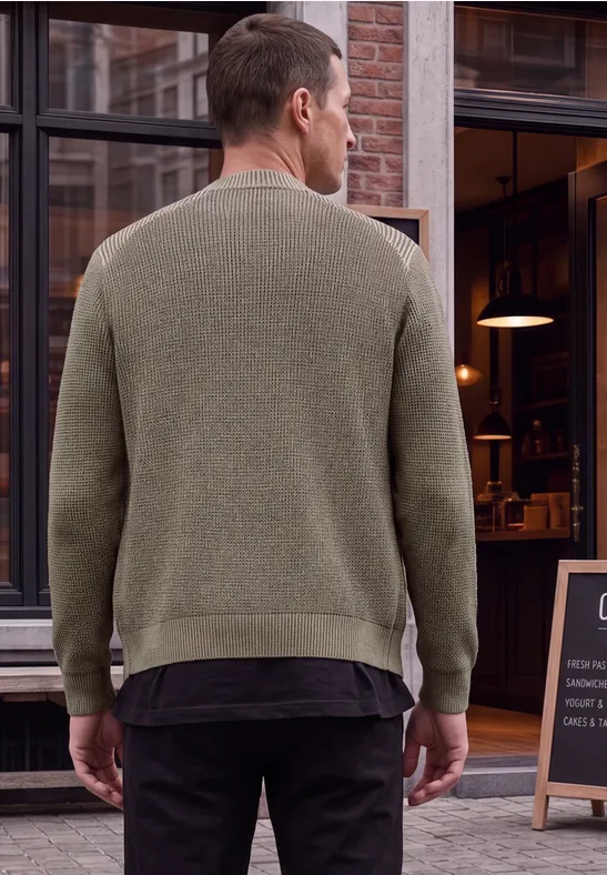 Ein Mann steht mit dem Rücken zur Kamera und trägt einen olivgrünen Pullover vor einem Schaufenster.