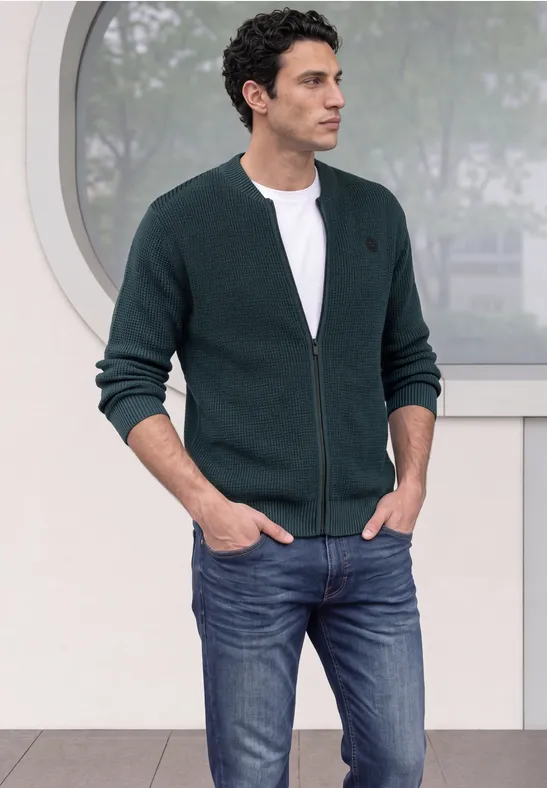 Ein Mann trägt eine dunkelgrüne Strickjacke mit Reißverschluss, weißes T-Shirt und Jeans vor einem runden Fenster.