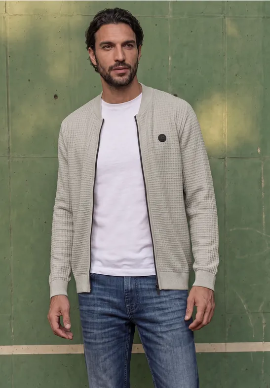 Ein Mann in heller Strickjacke und Jeans steht lässig vor einer grünen Wand.
