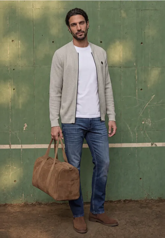 Ein Mann trägt Jeans, weißes T-Shirt, graue Jacke und braune Wildlederstiefel, hält Reisetasche vor grünem Hintergrund.