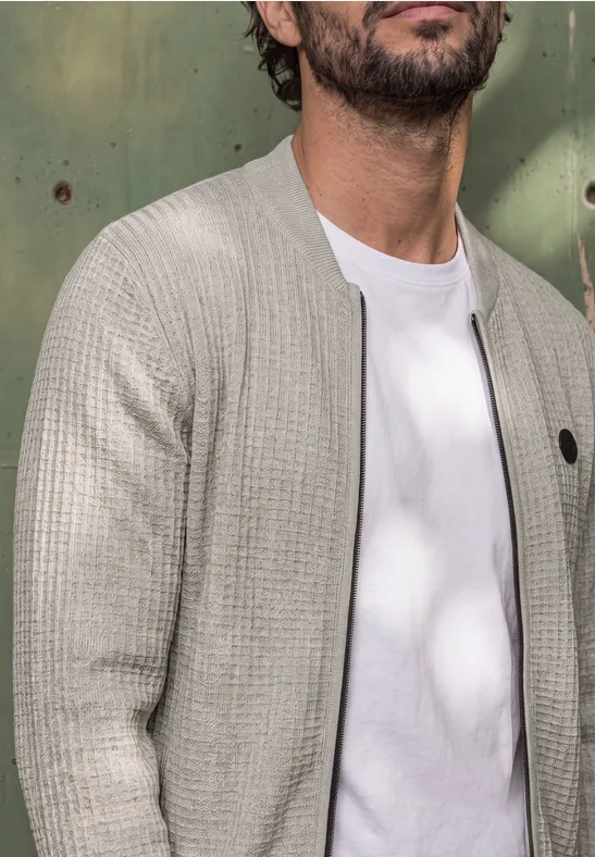 Ein Mann trägt ein weißes T-Shirt unter hellgrauem Strickcardigan vor grünem Hintergrund.