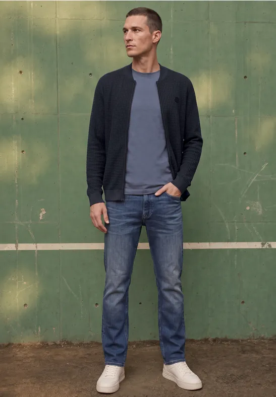Junger Mann in Jeans, grauem T-Shirt und dunkler Jacke steht vor grüner Wand.