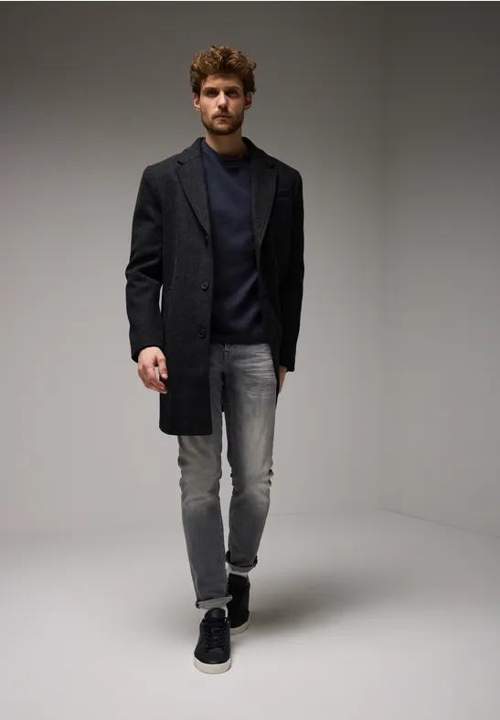 Mann in dunklem Mantel, Jeans und Sneakers steht in minimalistischem Raum.
