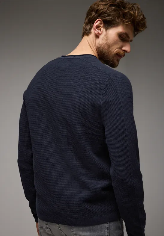 Mann mit braunen Haaren trägt dunkelblauen Pullover, steht mit dem Rücken zur Kamera.