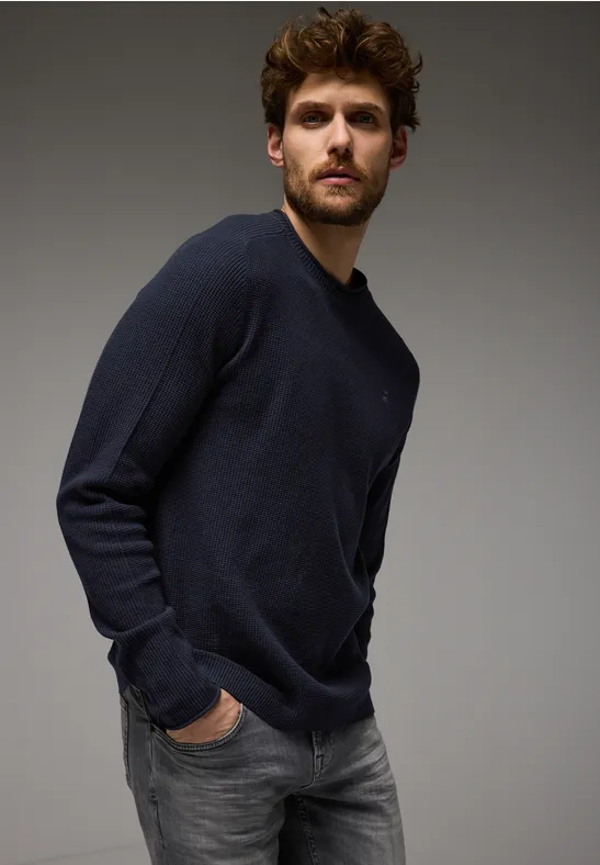 Mann mit dunkelblauem Pullover und grauer Jeans steht vor grauem Hintergrund.