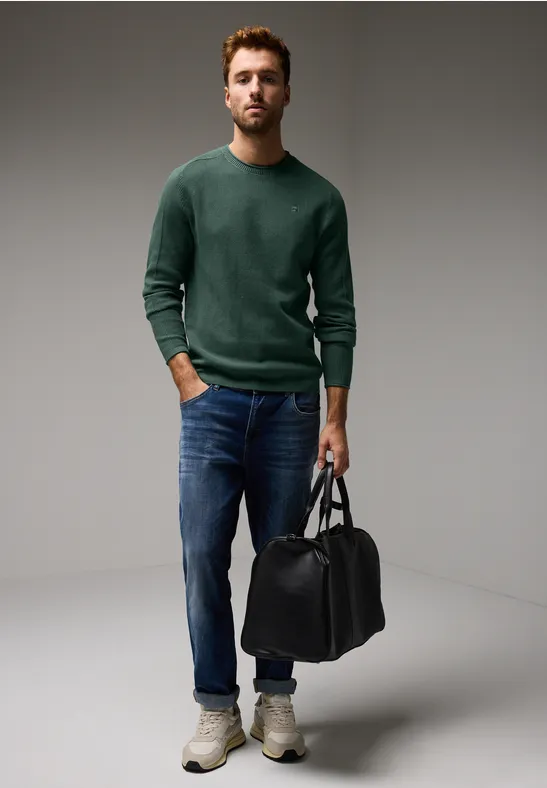 Mann mit grünem Pullover, Jeans und schwarzer Tasche steht im Studio.