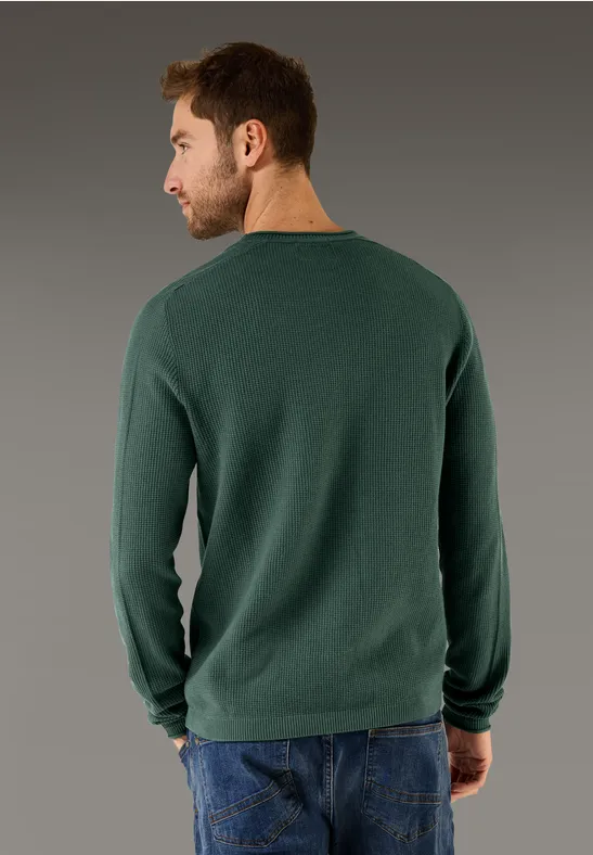 Mann mit grünem Pullover und Jeans von hinten vor grauem Hintergrund.