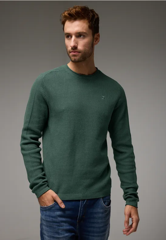 Ein Mann trägt einen grünen Pullover und Jeans, steht vor grauem Hintergrund.