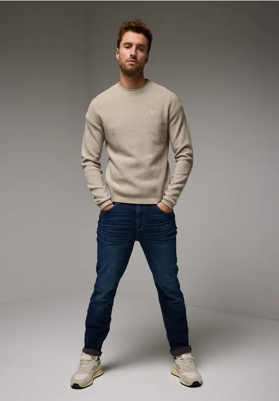Mann in beigem Pullover, blauer Jeans und Sneakers steht mit Händen in den Taschen.