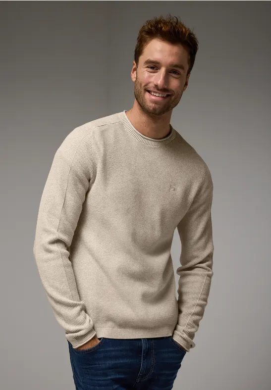 Mann mit hellem Pullover und Jeans lächelt in die Kamera.
