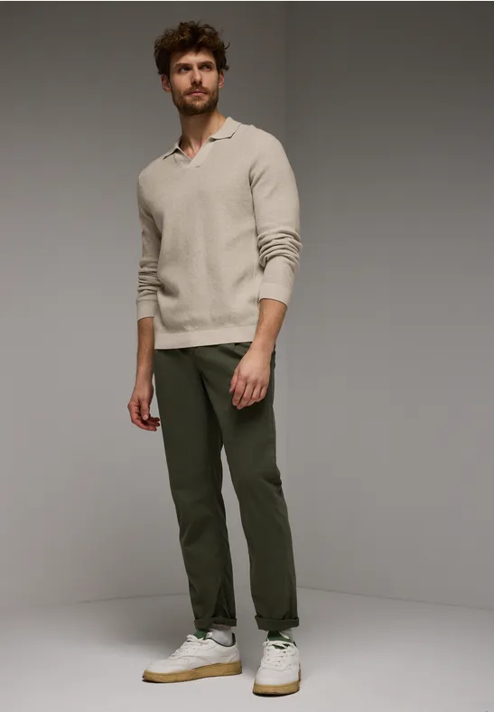Mann in beigem Pullover, grüner Hose und weißen Sneakers, steht vor grauem Hintergrund.