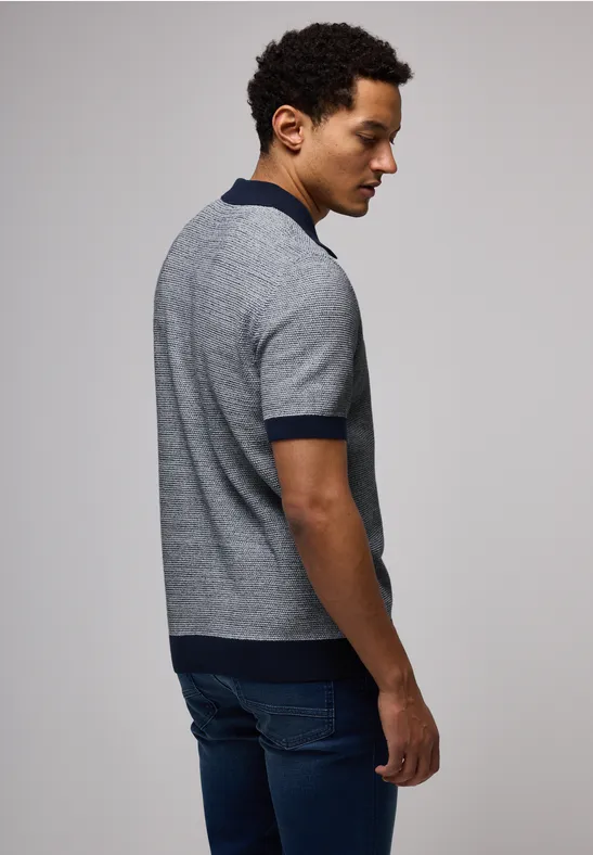 Mann mit kurzem Haar trägt graues Shirt und Jeans, steht seitlich und blickt nach unten.