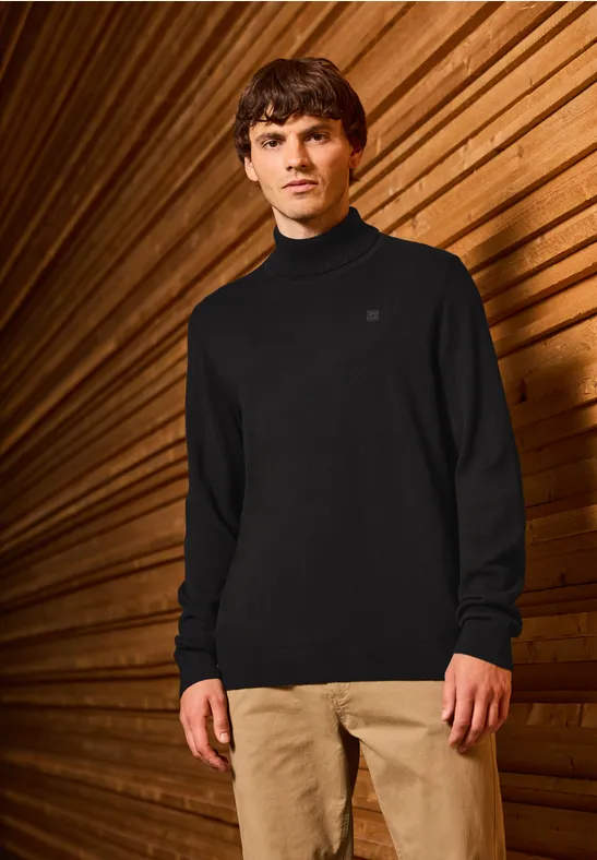 Mann in schwarzem Pullover und beiger Hose vor Holz-Wand.