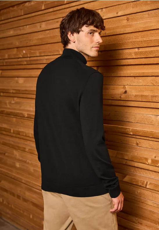 Mann in schwarzem Pullover vor Holzwand, sieht über die Schulter zurück.