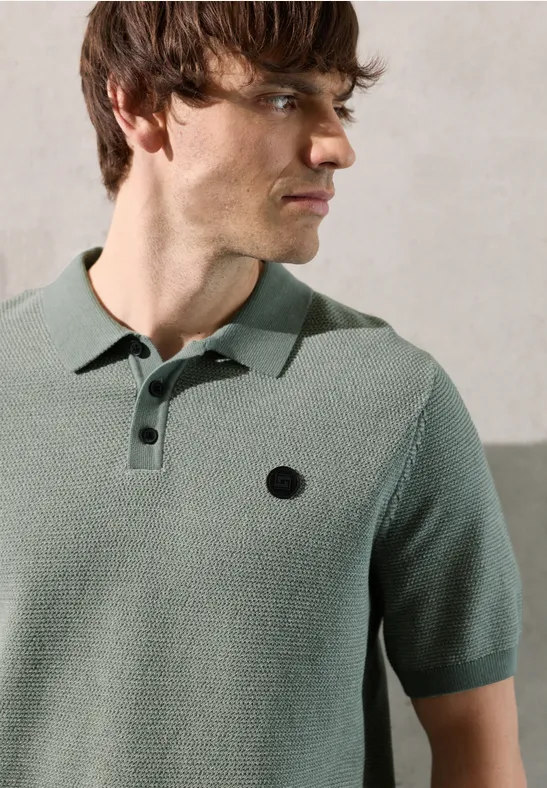 Een man draagt een lichtgroen polohemd met korte mouwen en een zwart logo op de borst.