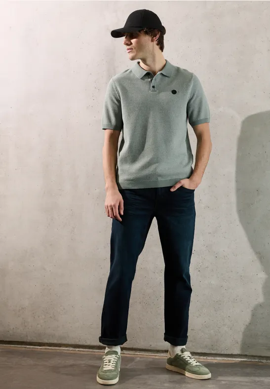 Jonge man met grijs poloshirt, spijkerbroek, groene sneakers en zwarte pet tegen muur, handen in zakken.