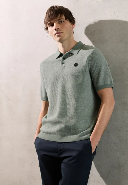 Een man leunt tegen een muur, draagt een lichtgroen poloshirt en een donkere broek, handen in de zakken.