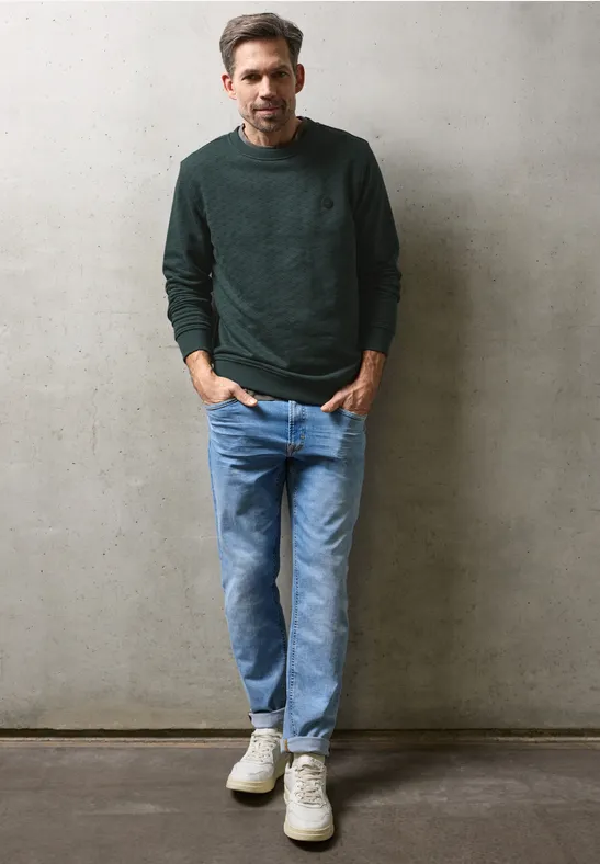 Ein Mann in grünem Pullover und blauen Jeans lehnt lässig vor einer grauen Wand.