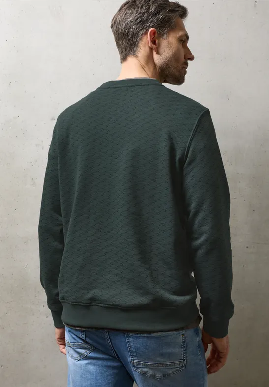 Ein Mann von hinten in grünem Stepppullover und Jeans vor hellem Hintergrund.
