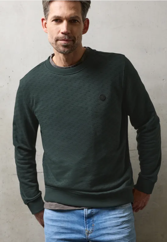 Ein Mann trägt einen dunkelgrünen, strukturierten Pullover und helle Jeans vor einer schlichten Wand.