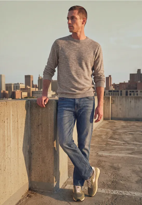 Ein junger Mann lehnt lässig an einer Betonmauer auf einem Parkhausdach, mit Skyline im Hintergrund.