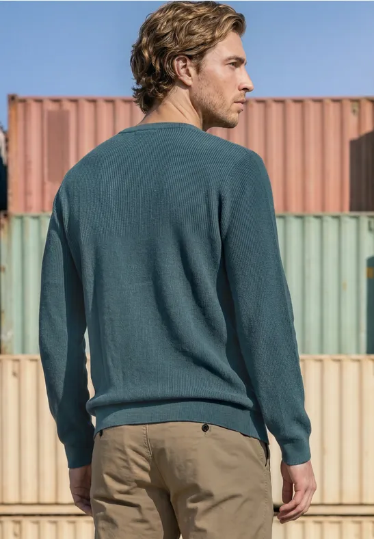 Un homme en pull vert et pantalon beige, vu de dos, se tient devant des conteneurs colorés.