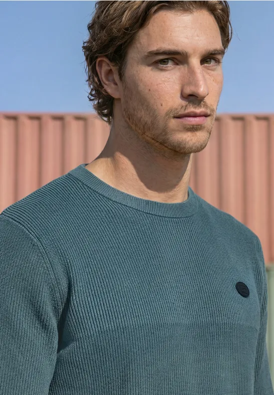 Portrait d’un jeune homme en pull turquoise devant un container rose, regard pensif.