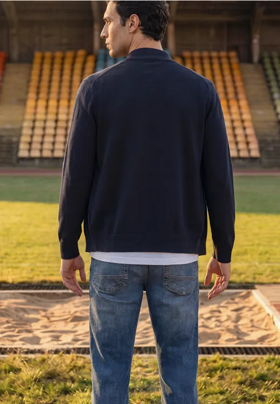 Een man van achteren in spijkerbroek en donker shirt bij een zandbak op een atletiekbaan in een stadion.