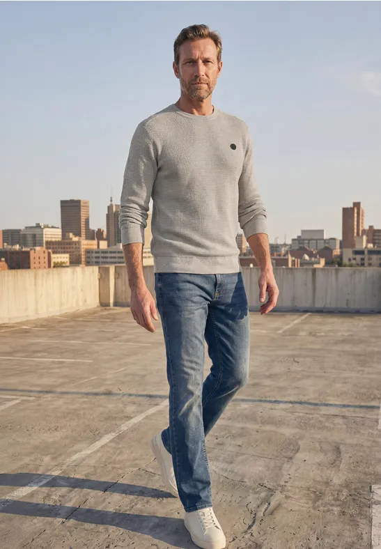 Ein Mann in Jeans und Pullover steht auf einem Dach mit urbaner Skyline im Hintergrund.
