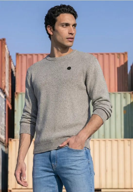 Een man in een grijze trui en blauwe jeans staat voor kleurrijke containers in de buitenlucht.